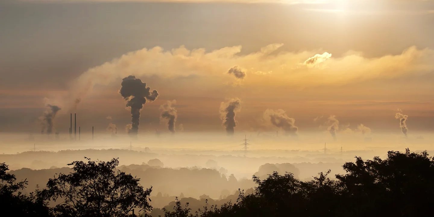 Industrial skyline with tall smokestacks releasing smoke at sunset over a hazy landscape