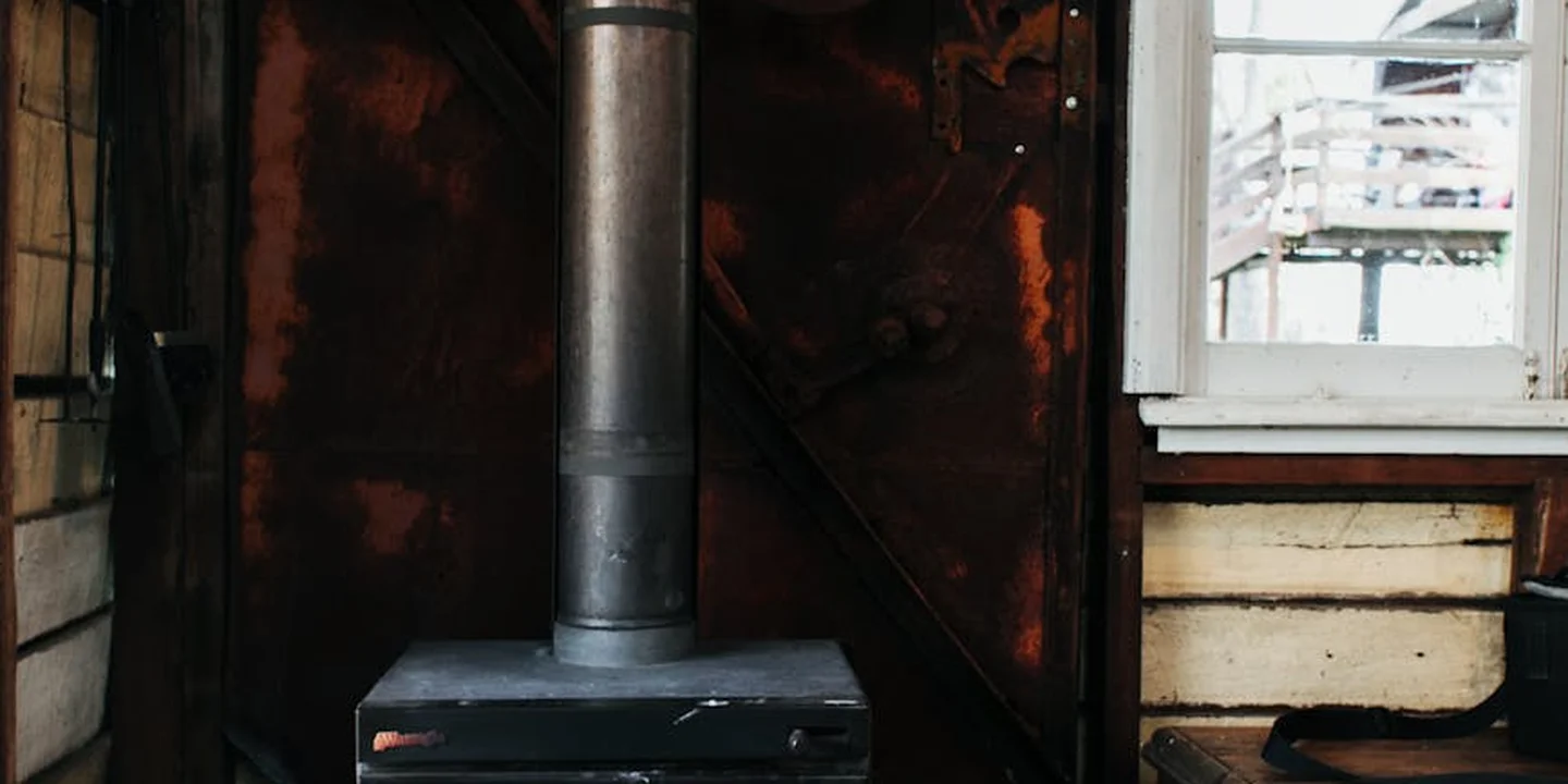 Metal chimney flue extending through a dark attic with wooden siding and a window on the right.