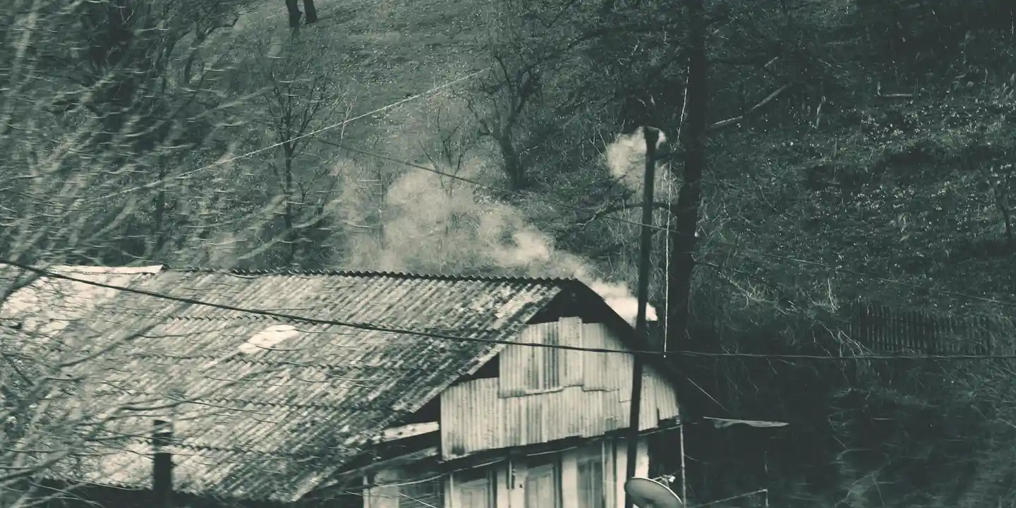 Black-and-white photo of a weathered house with smoke rising from a chimney in a smoky, wooded setting.