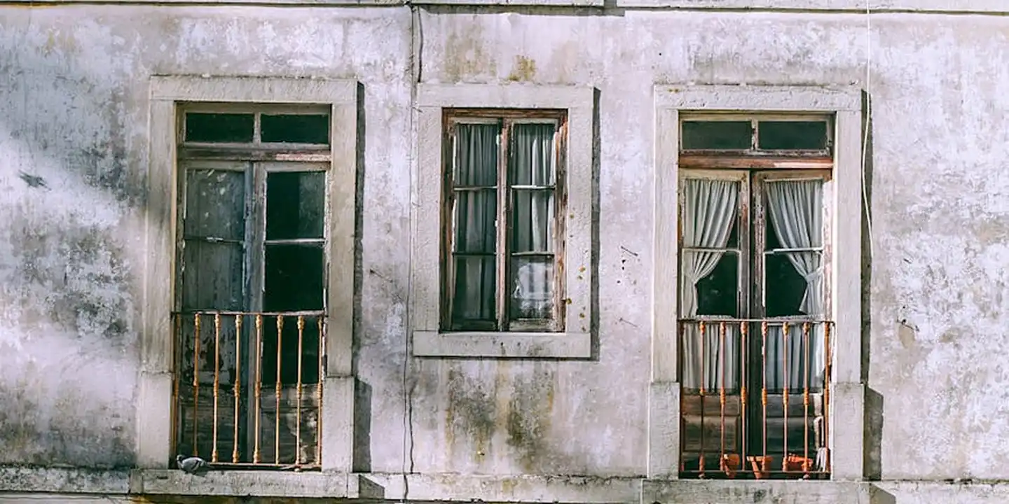 Weathered white building facade with three window openings, some with curtains and metal bars, representing older masonry structures.