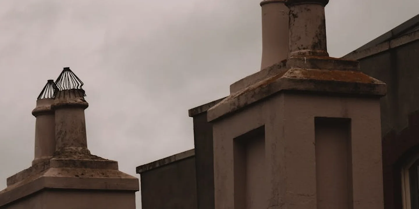Two brick chimneys on a sloped roof under a cloudy sky; the left chimney has a metal cage cap, while the right chimney has a solid cap.