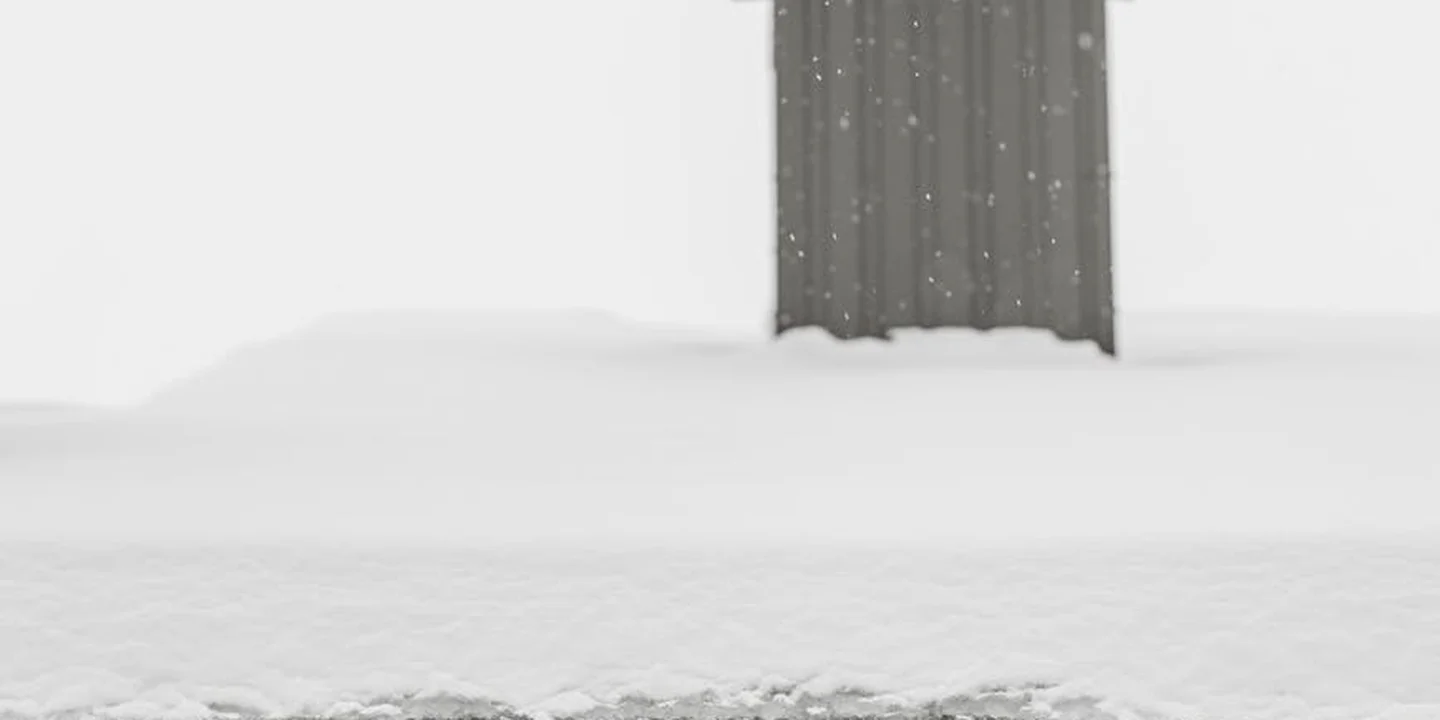 Snow-covered exterior wall with a dark metal vent/door protruding above the snow