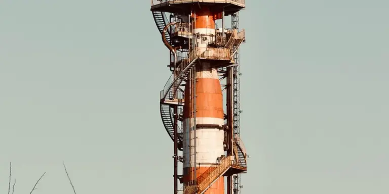Tall brick chimney wrapped in orange and white scaffolding with a metal staircase against a pale sky.