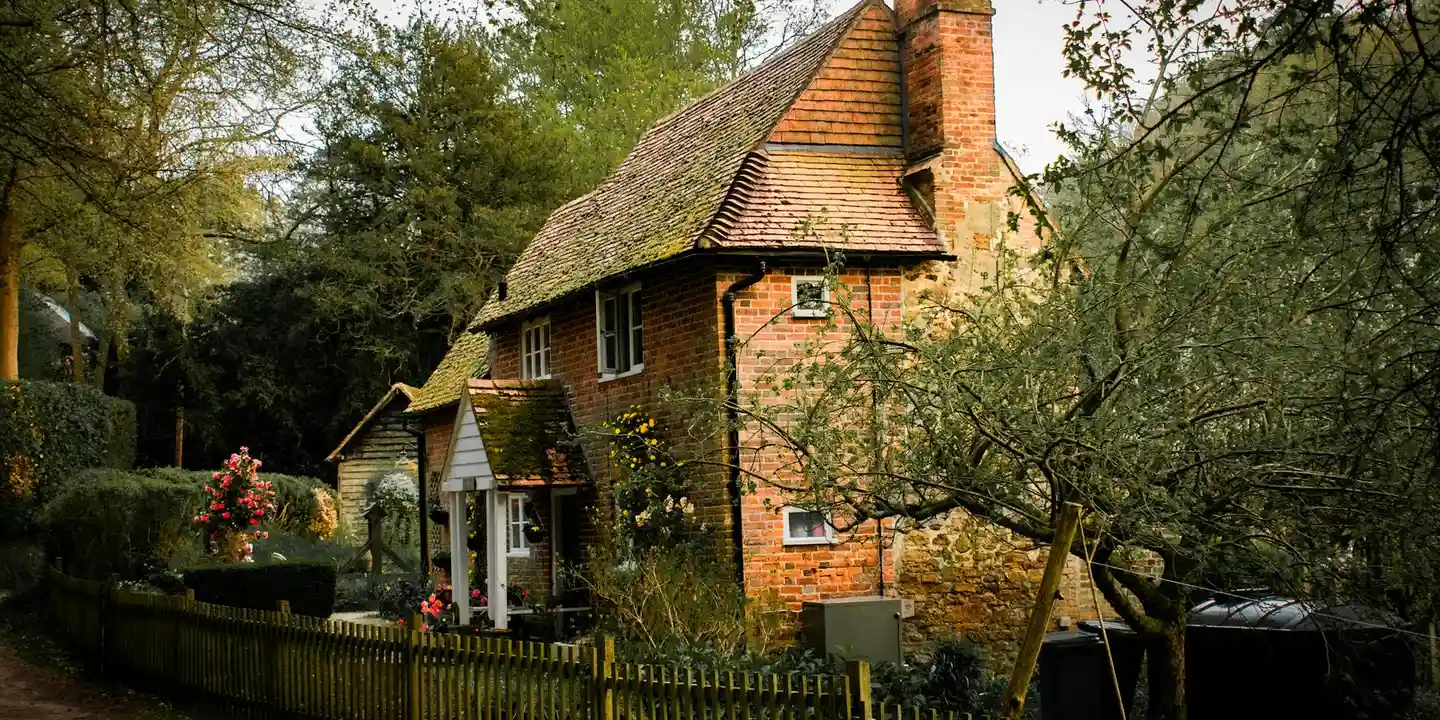 Brick cottage with a tall chimney on a steep roof, surrounded by trees.