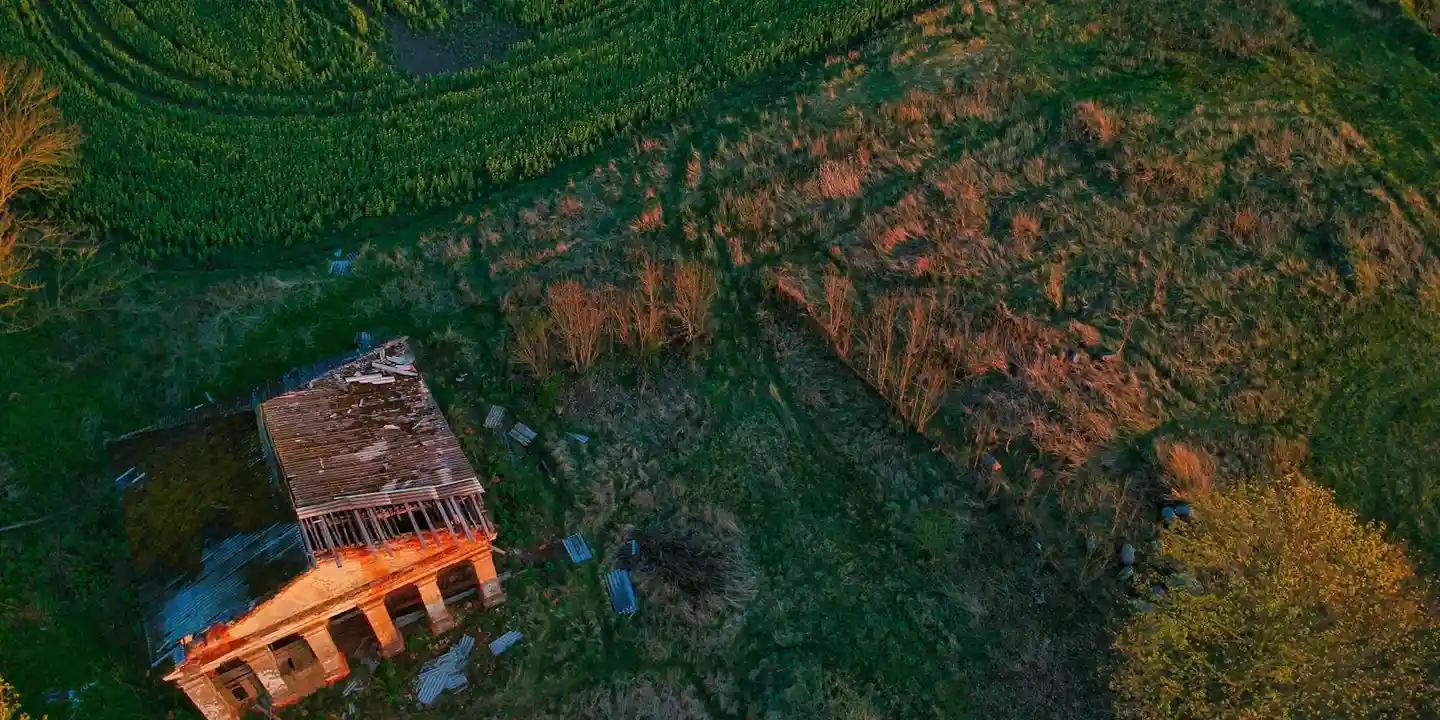 Aerial view of a small building with a damaged roof in a wooded hillside area, showing exposed framing and scattered debris.