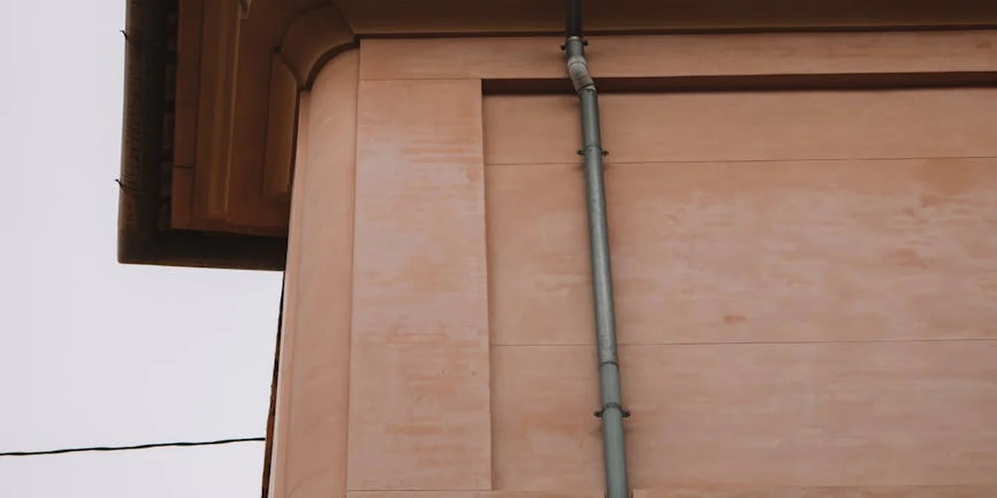 Close-up of a house gutter and vertical downspout mounted on a peach-toned wall, illustrating a typical drainage setup.
