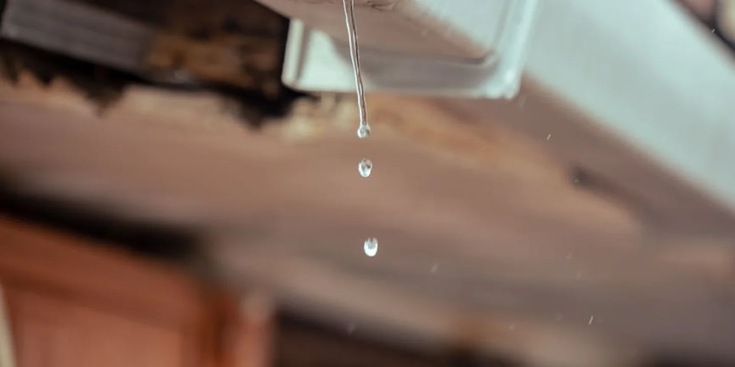 Close-up image of a metal gutter edge with water droplets dripping, with a blurred wooden background.