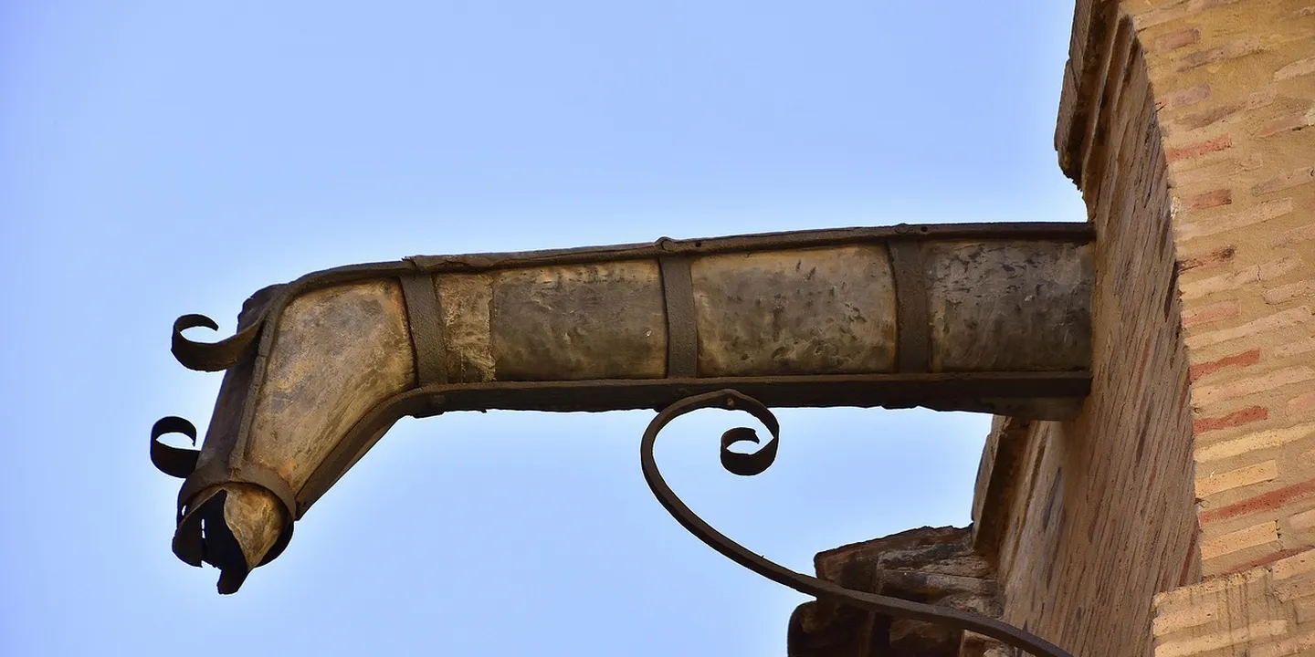 Close-up of a weathered gutter on a brick house with a curved bracket and downspout against a blue sky.