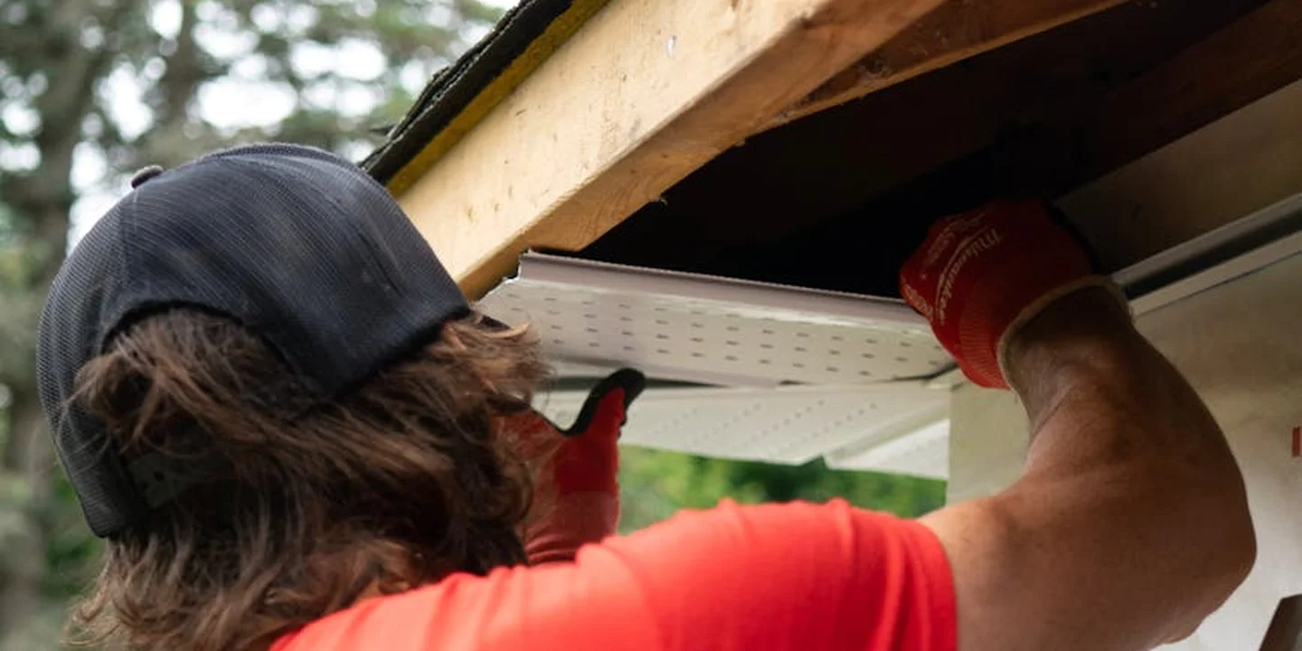 Person wearing a dark cap and red shirt installs a white gutter under a roof edge, wearing gloves.