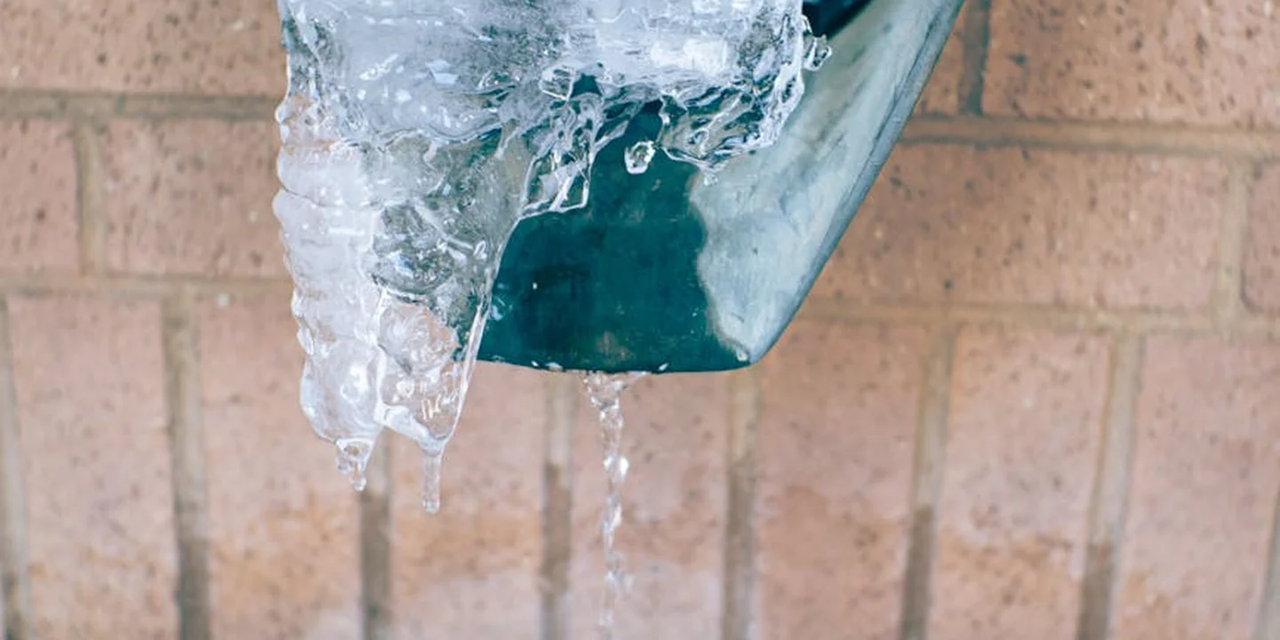 Close-up of a damaged green gutter with water leakage and ice along the edge, mounted over a brick wall.