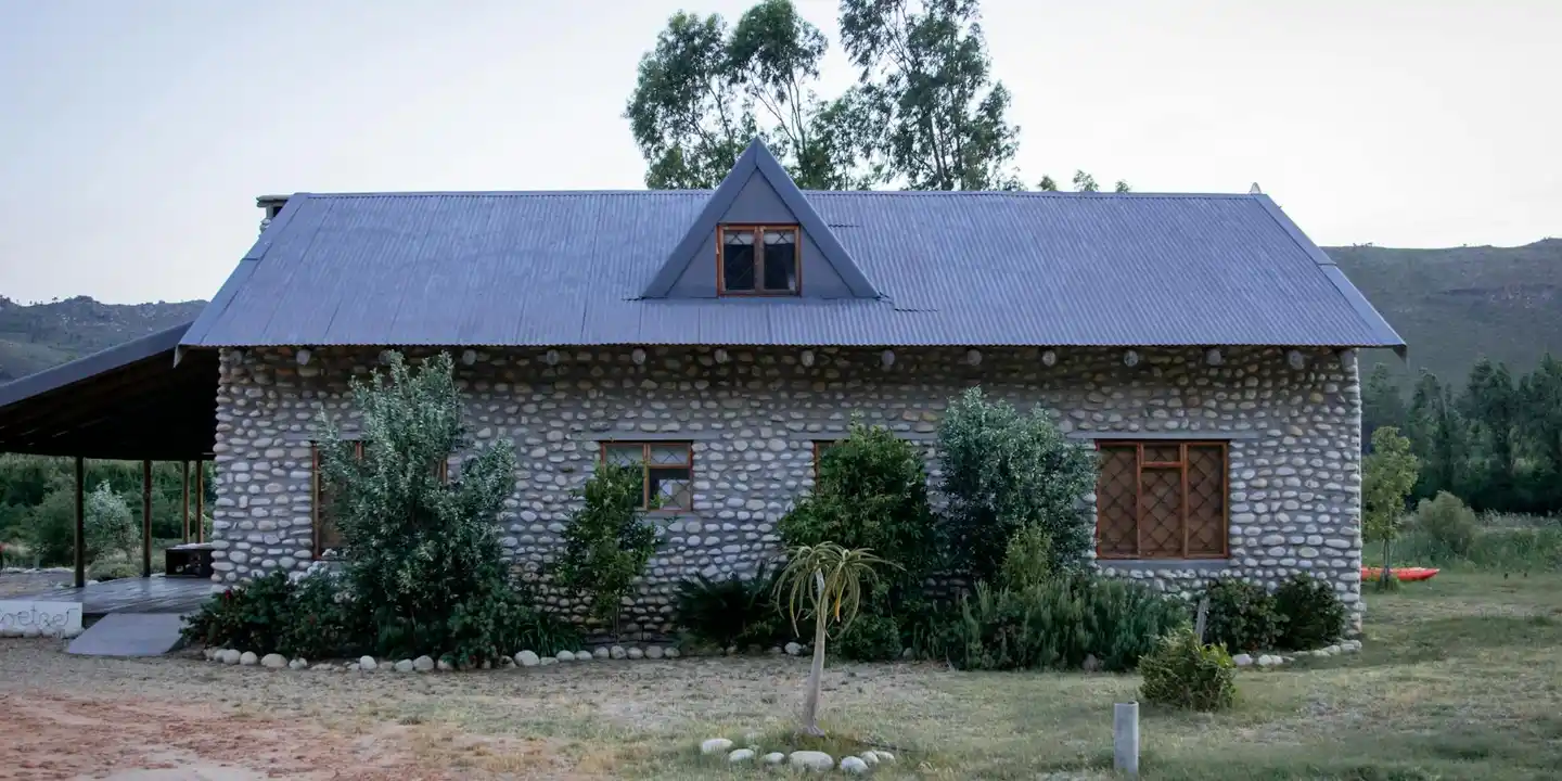 Rural stone house with a metal pitched roof and surrounding shrubs; no active construction visible.