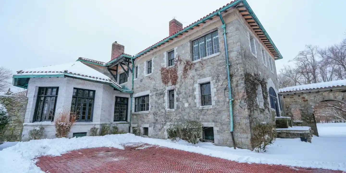 Snow-covered two-story house with a stone and stucco exterior, bare trees, and snow on the roof, illustrating winter conditions that can cause ice dams.