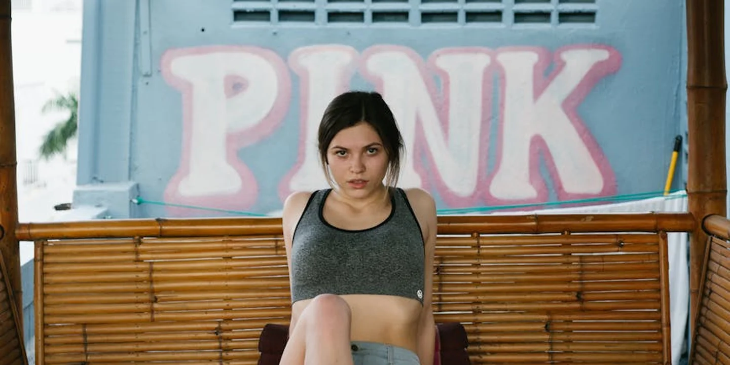 Young woman with dark hair wearing a gray crop top seated on a wooden bench in front of a blue wall with a large pink sign.