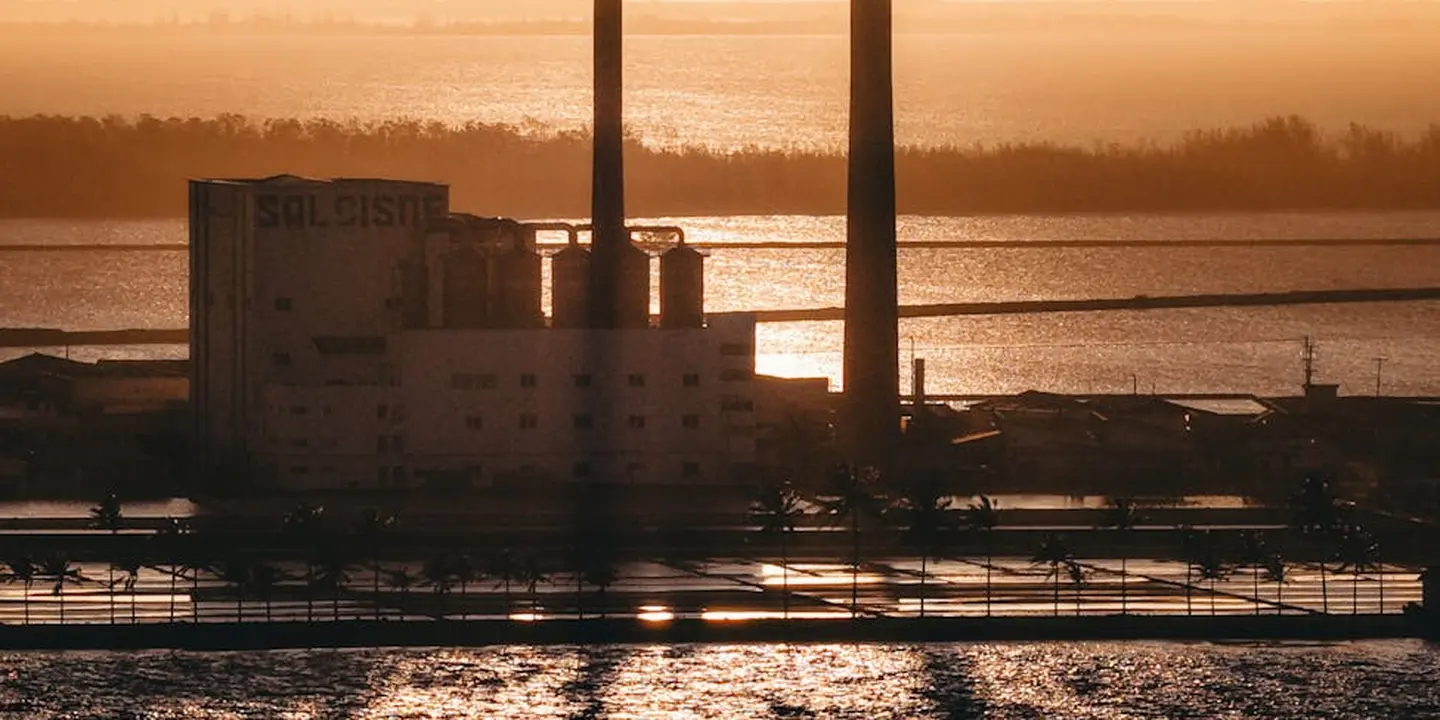 Industrial smokestacks silhouetted at sunset over a waterfront facility, with railings and equipment in the foreground.