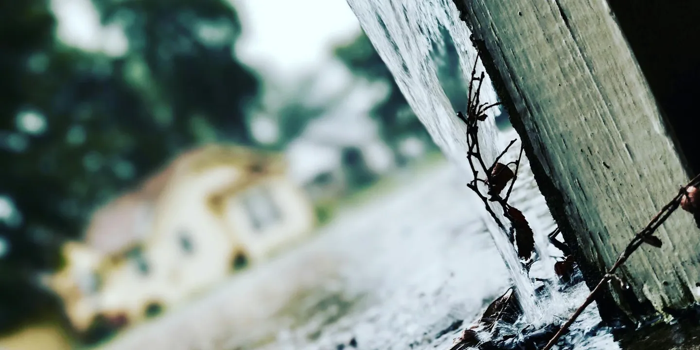 Close-up of a weathered wooden post with barbed wire and raindrops, with a blurred house and greenery in the background.