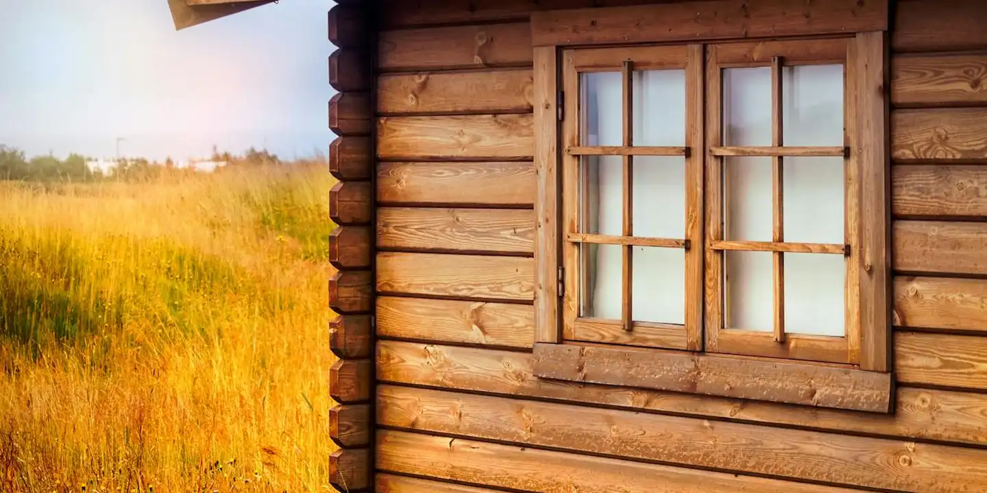 A rustic log cabin with a paneled window sits in a sunlit field with tall yellow grass under a clear sky.