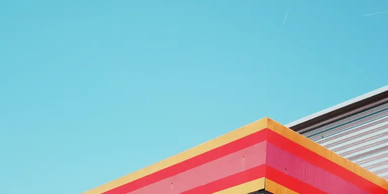 Close-up of a roof edge with stacked multi-colored fascia panels (yellow, orange, pink) against a clear blue sky.