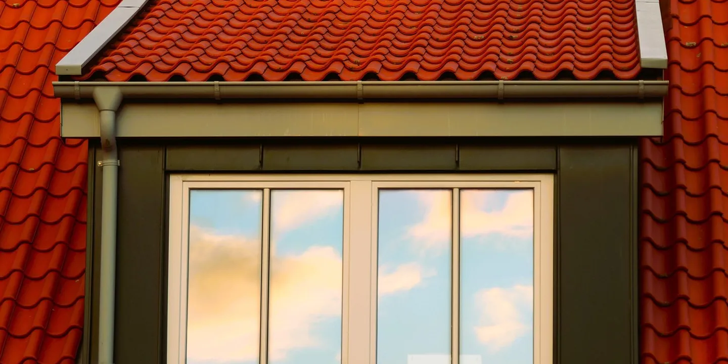 Close-up of a red-tiled roof with a white gutter along the eave, and a window below reflecting a blue sky.