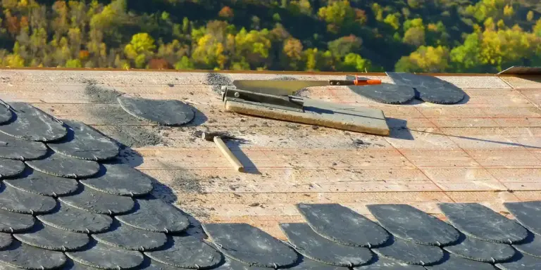 A sloped roof with exposed plywood decking and scattered slate shingles, with a few tools resting on the surface; autumn trees are visible in the background.