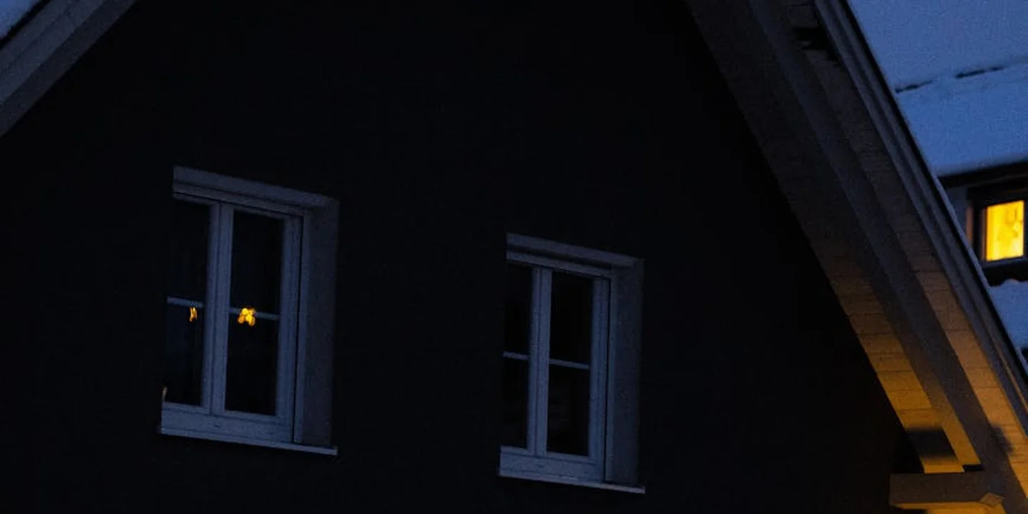 Exterior of a dark house at dusk with two white-framed windows and a warm-lit third window, showing the roof edge and eaves.