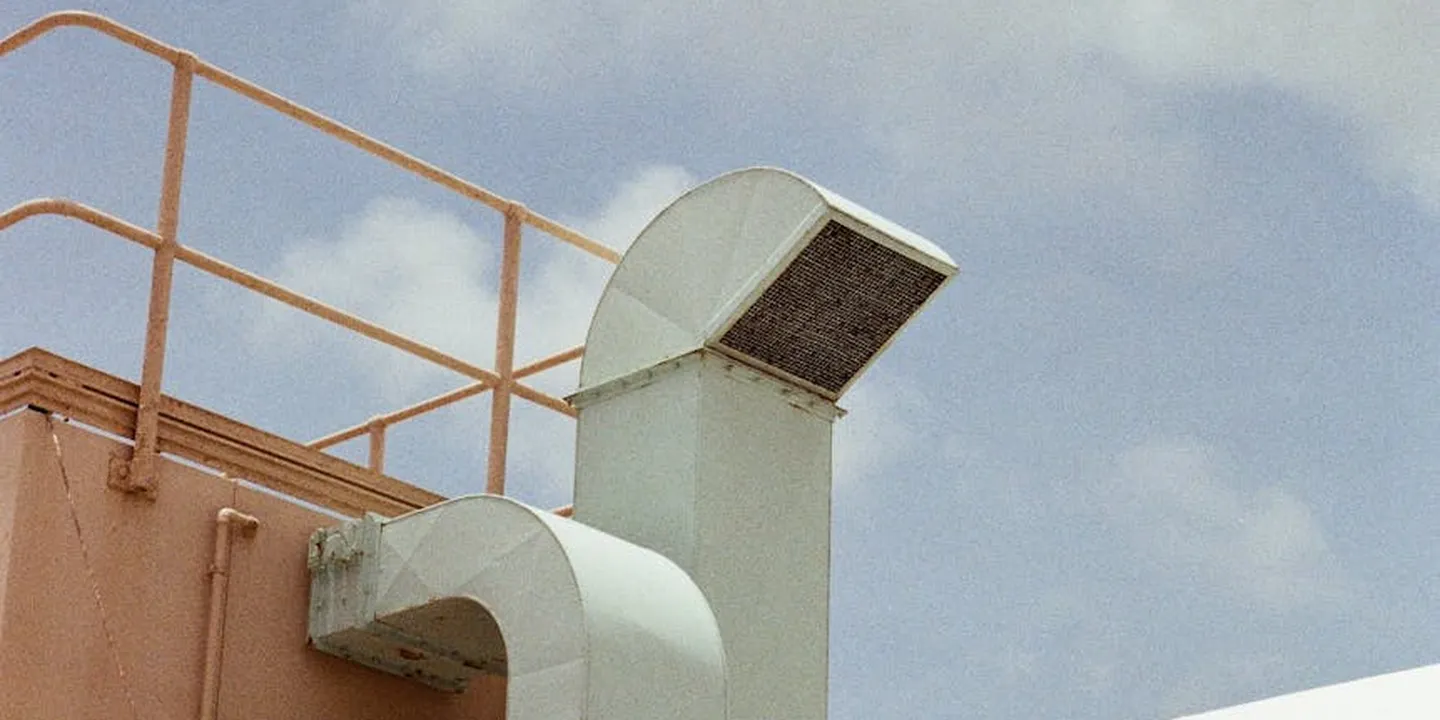 White metal roof exhaust vent protruding from a light orange roof with orange railing and a clear blue sky in the background.