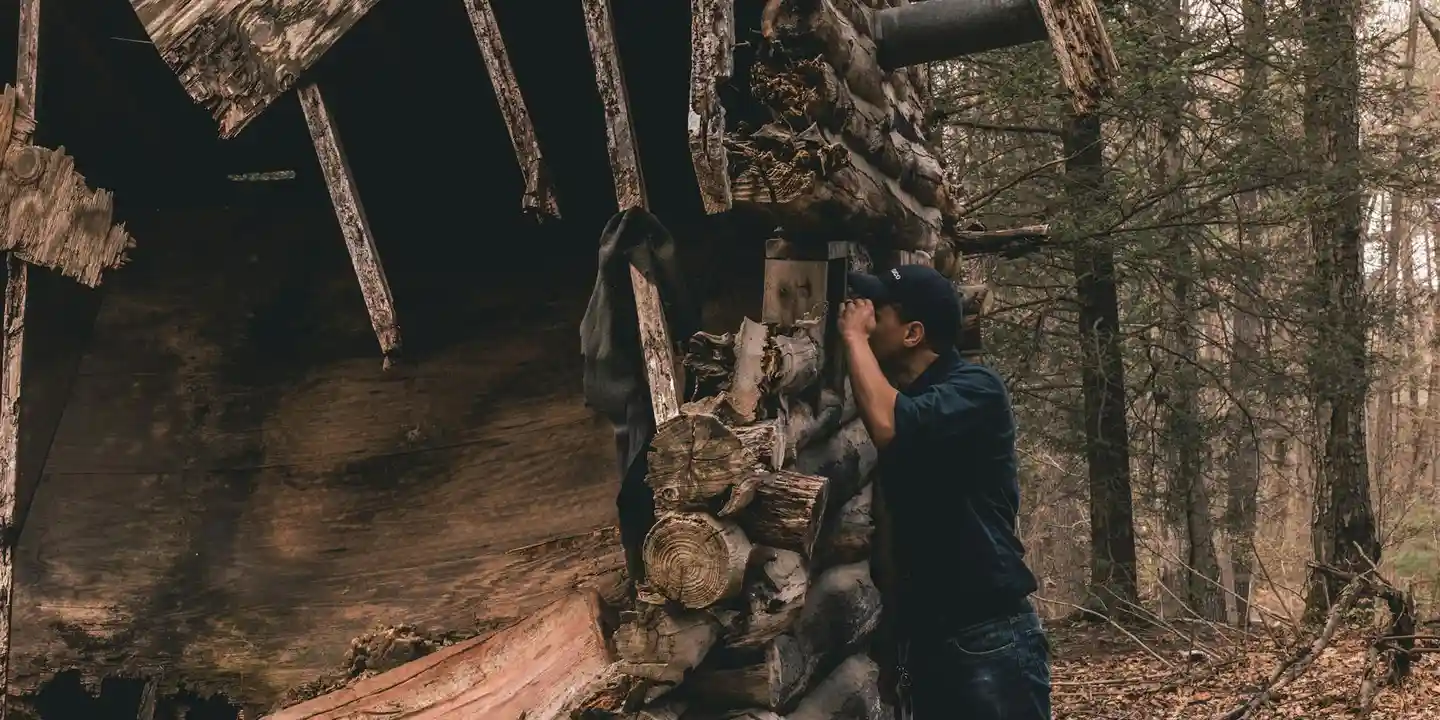 A person in dark clothing stands near a partially collapsed wooden shelter in a forest, with stacks of wood and debris around.