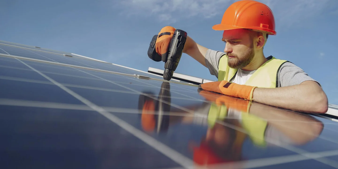 Solar installer wearing an orange hard hat and gloves tightens a component on solar panels mounted on a roof