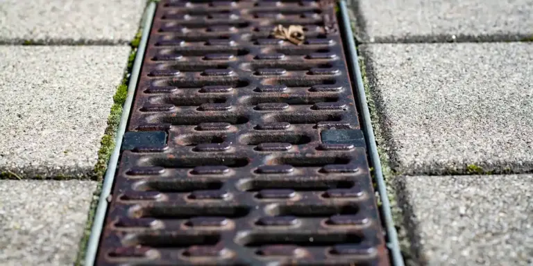 Close-up of a dark metal storm drain grate set in a gray concrete sidewalk with evenly spaced paver stones; a small leaf rests on the grate.