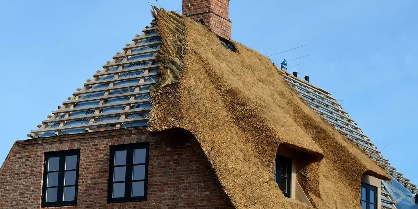 Brick house with a steep thatched roof under construction, scaffolding along the roof and a brick chimney visible.