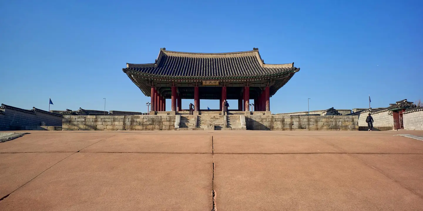 A traditional East Asian gate sits on a large stone terrace beneath a clear blue sky, with distant figures visible.